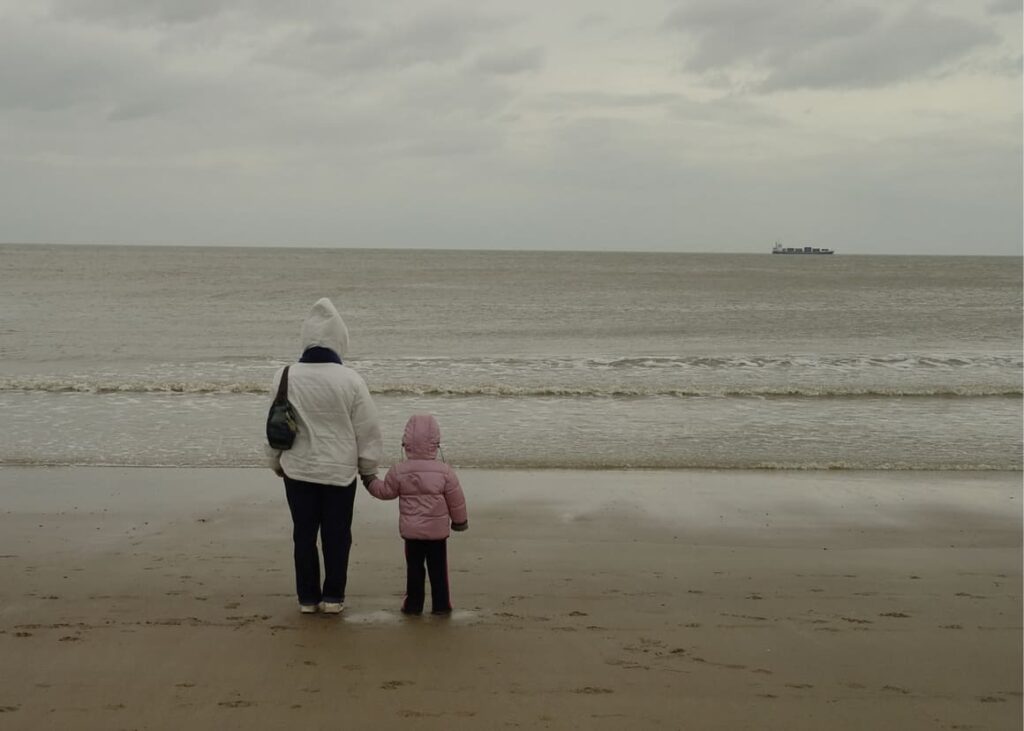A woman and child standing at the beach waving to a ship as they are leaving family behind