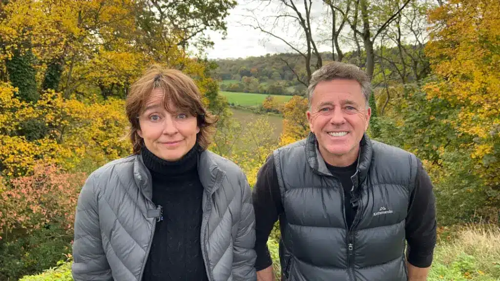 a husband and wife standing in front of a beautiful view in England recording a podcast about what it is like ot be back in England after sixteen years