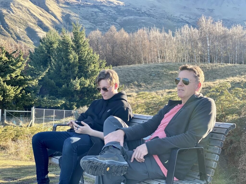 Father and son sitting on a bench. The son is looking at his phone